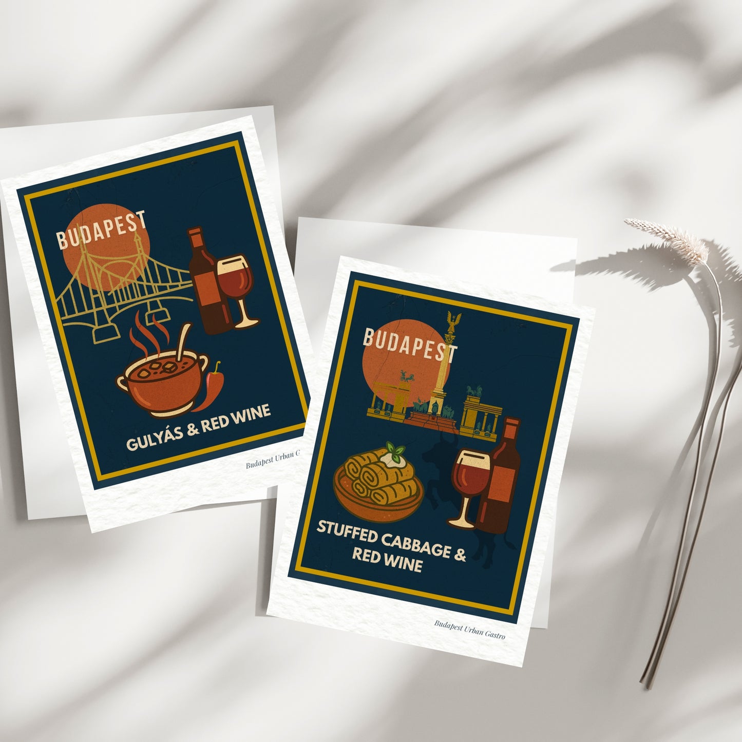 Two Budapest Urban Gastro navy postcards showing gulyás with red wine and stuffed cabbage with red wine, arranged on a bright minimal surface with soft natural shadows.