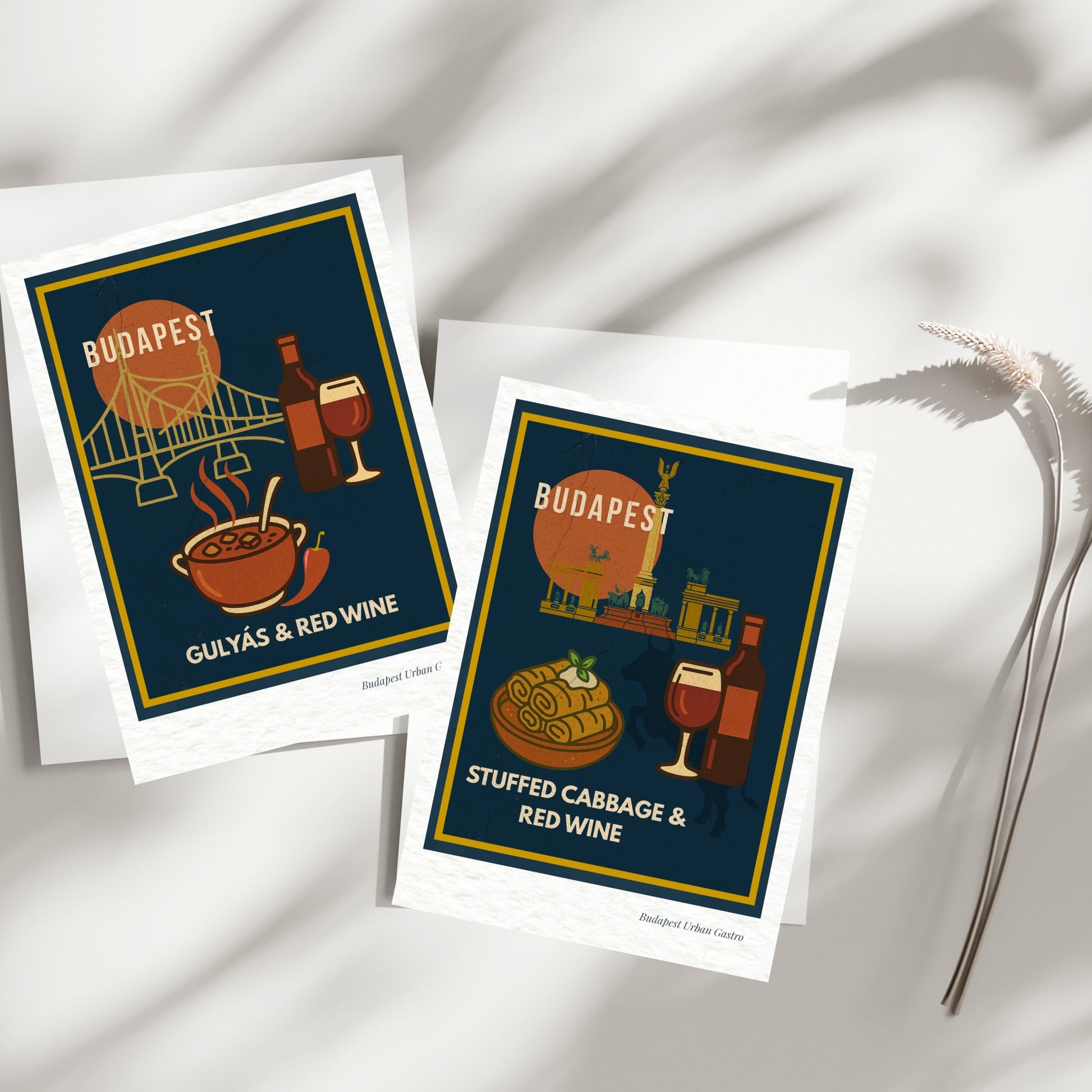 Two Budapest Urban Gastro navy postcards showing gulyás with red wine and stuffed cabbage with red wine, arranged on a bright minimal surface with soft natural shadows.