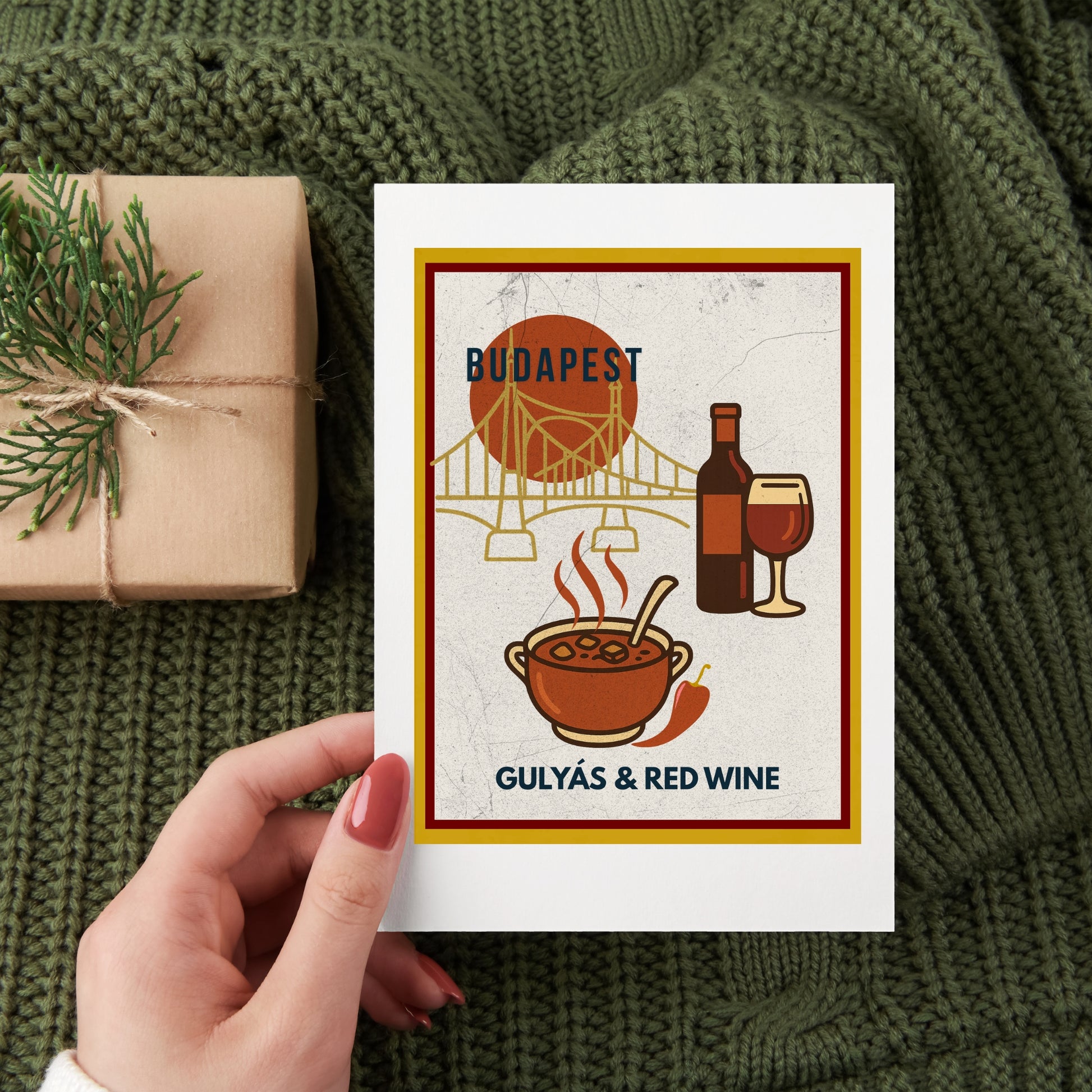 Hungarian food-themed art print featuring goulash and red wine, photographed as a flat lay with gift wrapping on a cozy textile background, suggesting a thoughtful travel-inspired gift.